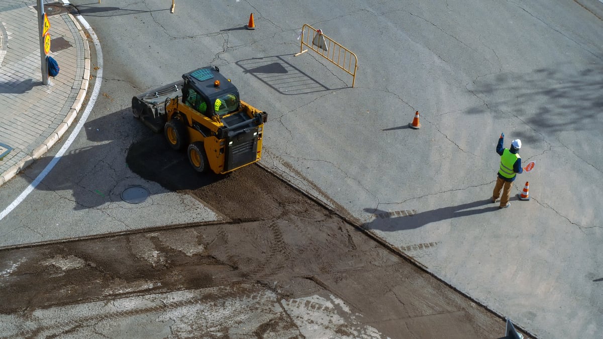 Construction worker guiding a skid steer loader paving a road section