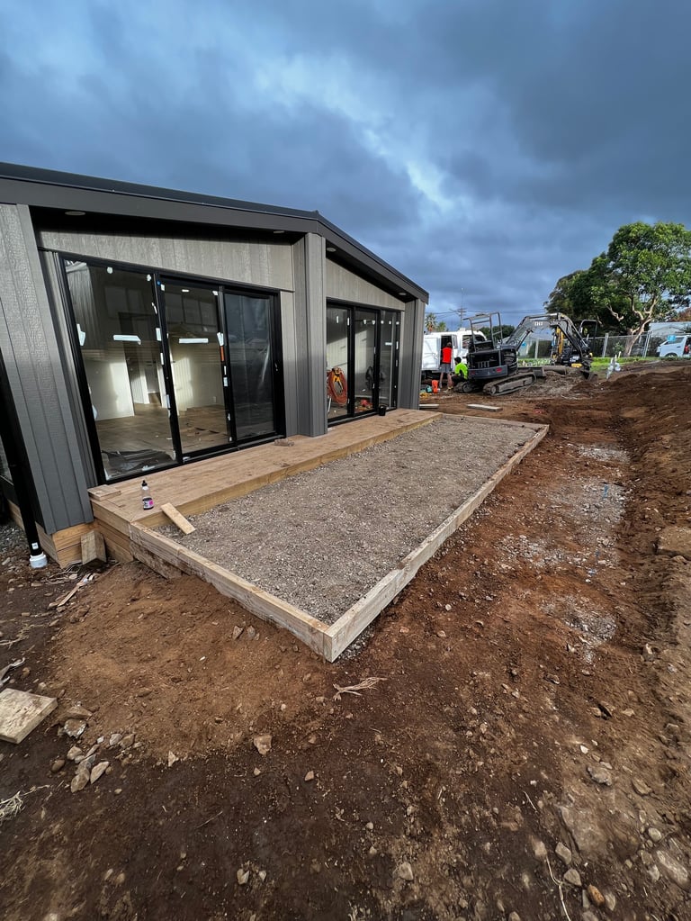 Modern prefab building with dark exterior under construction, featuring concrete foundation and gravel patio on a construction site