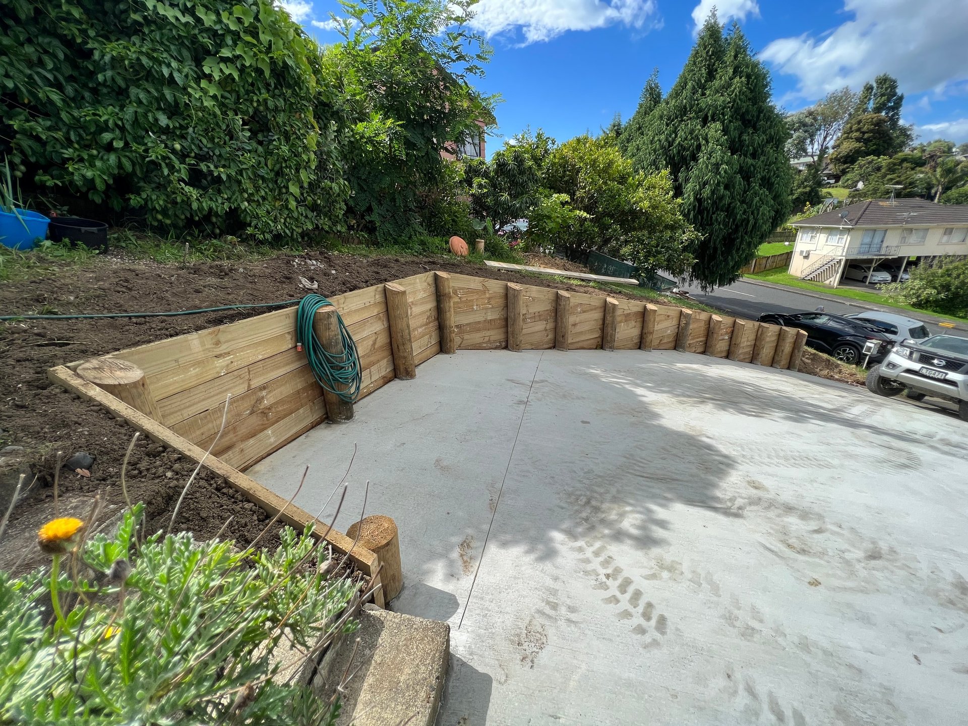 Curved wooden retaining wall with concrete base in a garden, surrounded by green landscaping and trees