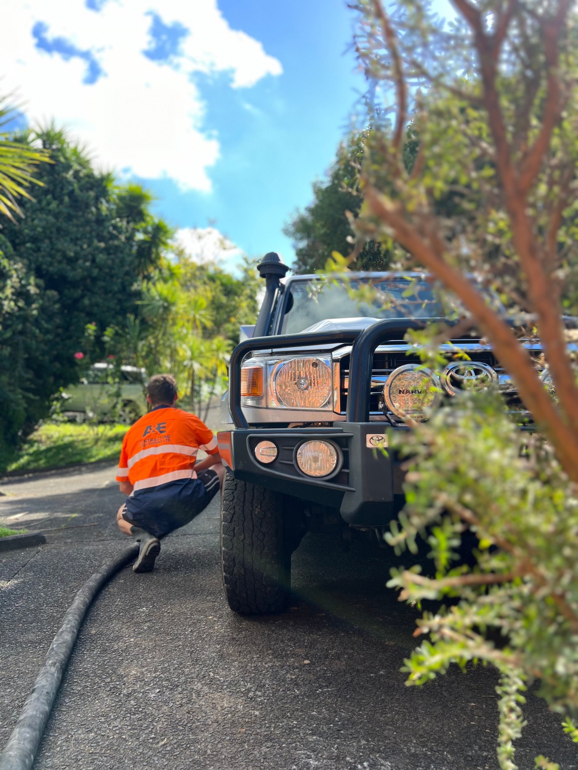 Person in orange safety vest crouching next to black SUV on tree-lined road with blue sky above