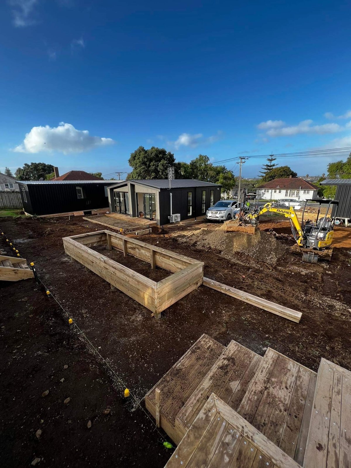 Construction site with wooden garden beds, black modular buildings, yellow excavator, and residential houses under a blue sky