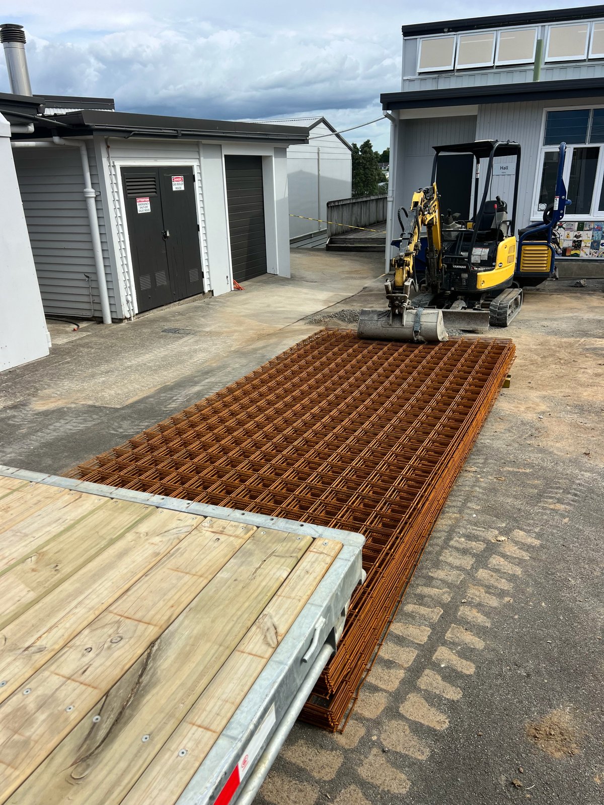 Construction site showing concrete foundation with rebar grid, wooden deck framing, yellow excavator, and residential buildings under development