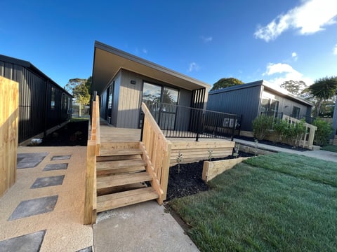 Modern prefab cabin with wooden deck and metal exterior, surrounded by green lawn and trees under blue sky