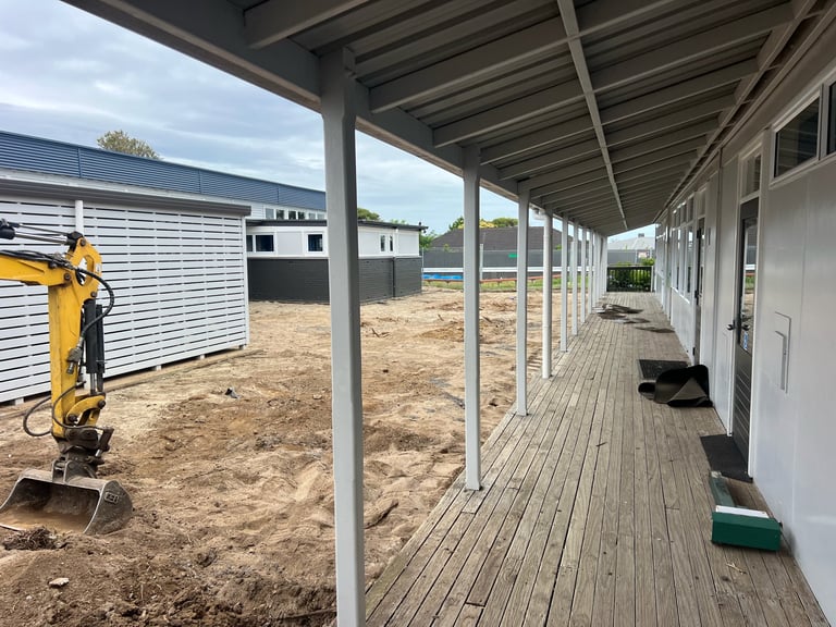 Covered walkway with wooden deck beside construction site with metal storage buildings and yellow machinery