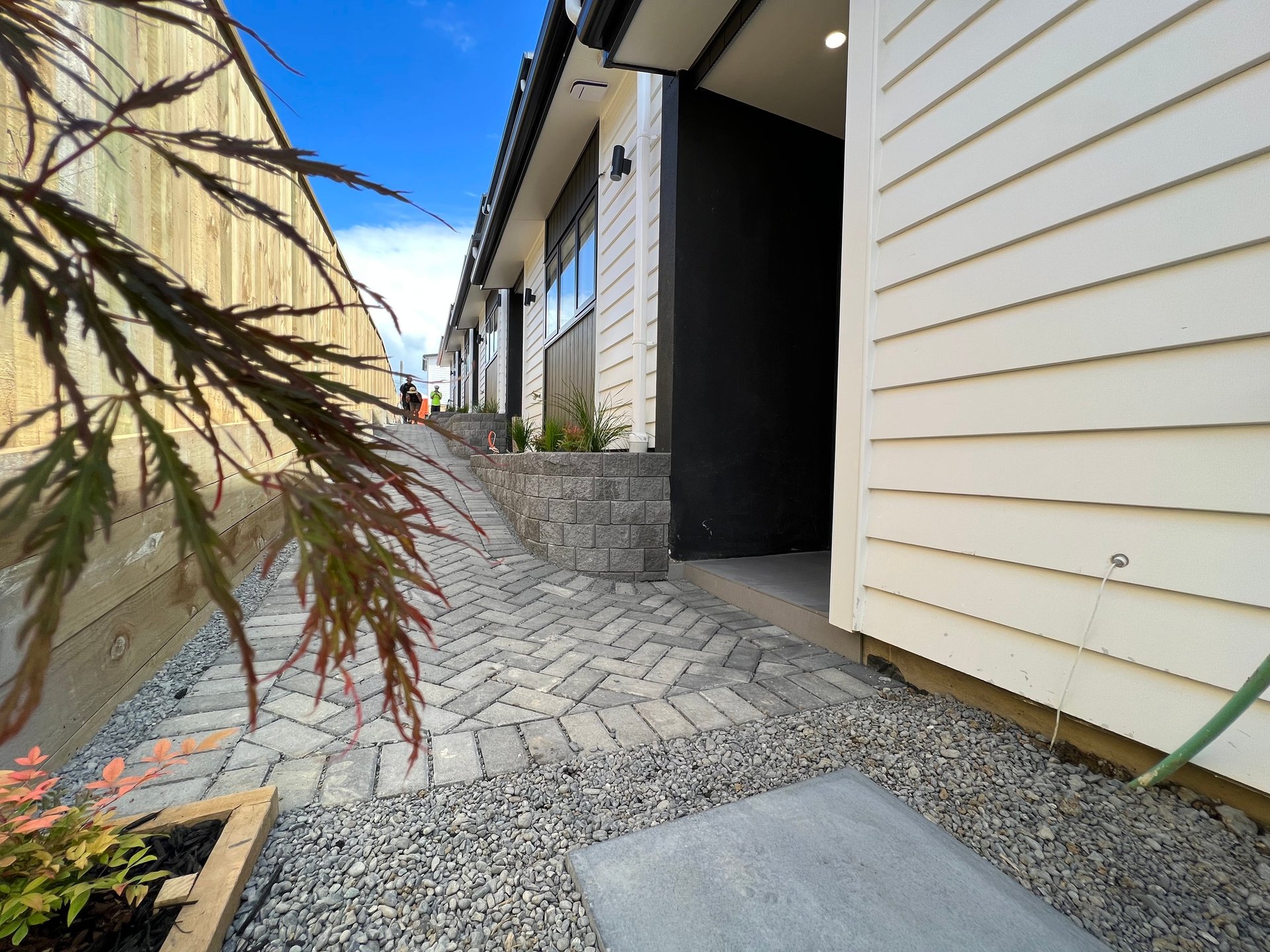 Modern residential home exterior with paved entryway, cream-colored siding, decorative gravel landscaping, and evergreen plants along the pathway