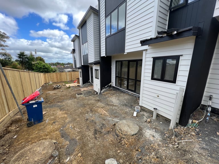 Modern black and white townhouses under construction with bare dirt yard, red wheelie bin, and blue recycling bin visible