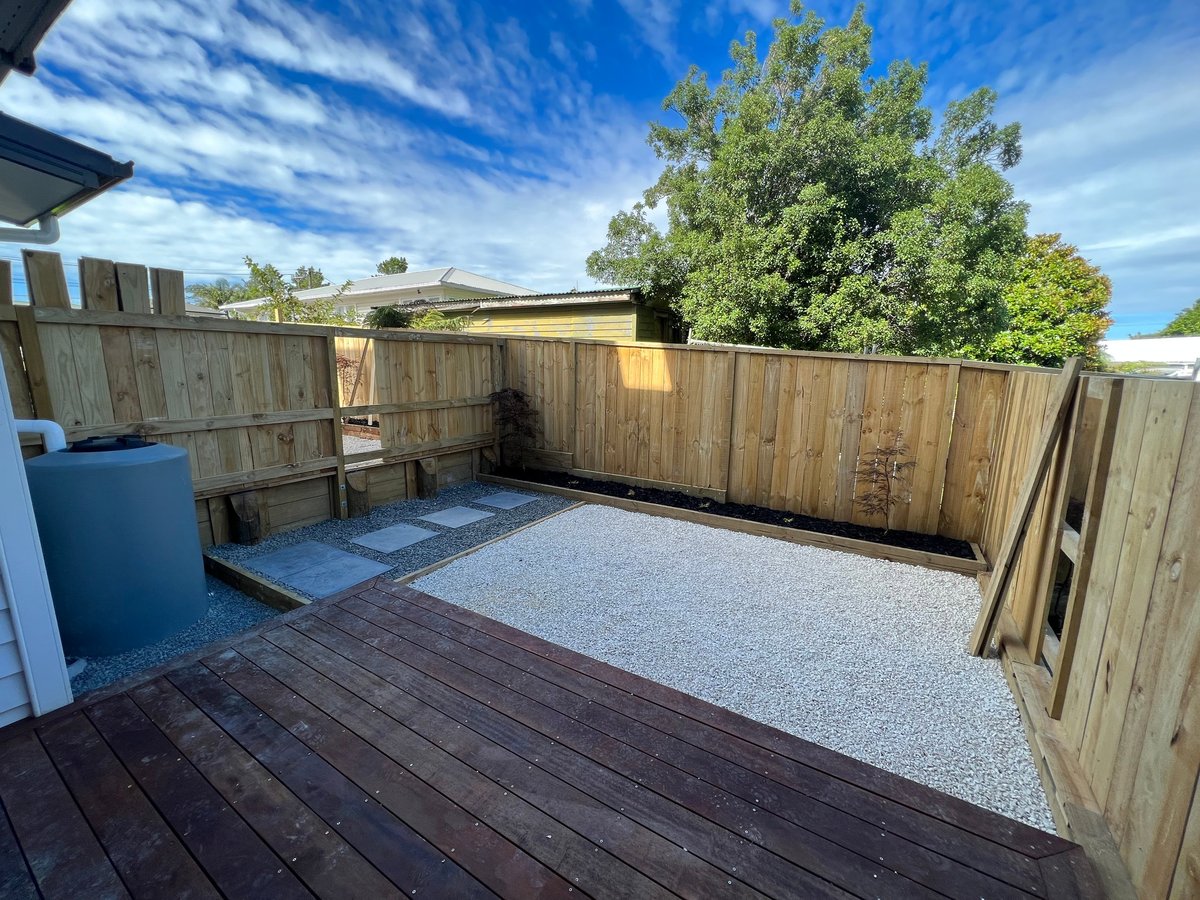 Empty backyard with wooden deck, gravel area, timber fence, and blue water tank under clear blue sky
