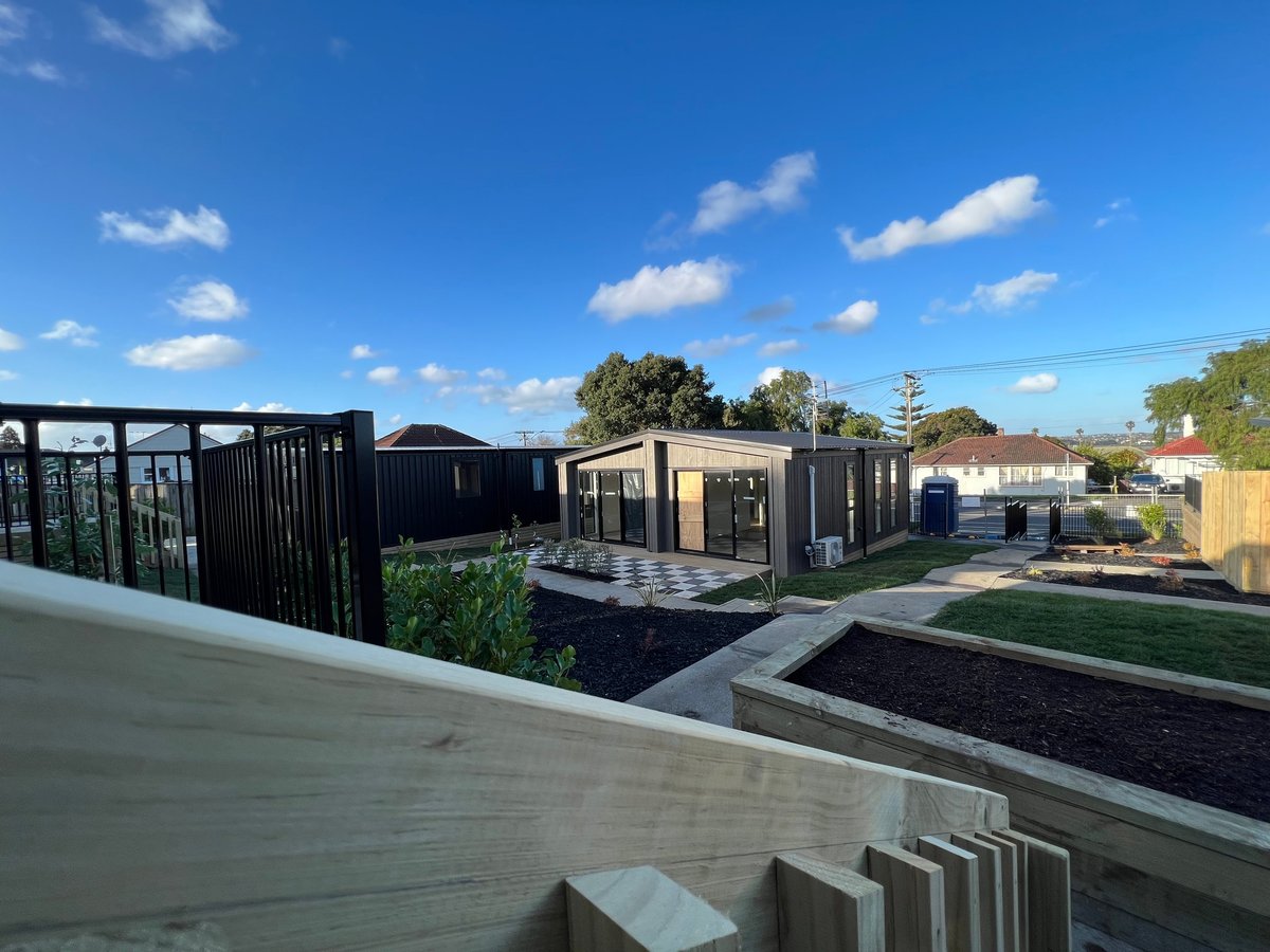 Elevated view of a modern residential compound with black metal fencing, beige and black modular buildings, manicured lawns, and clear blue sky with scattered clouds