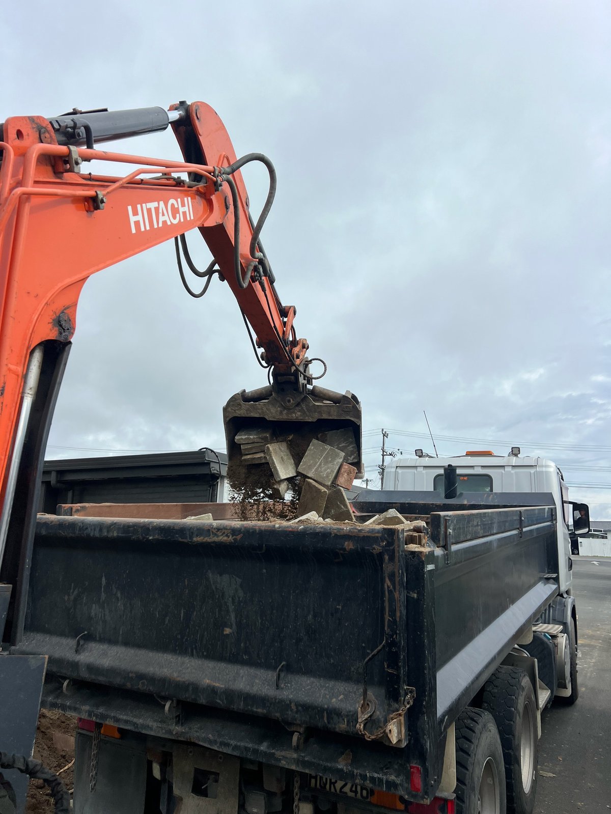 Orange Hitachi excavator with hydraulic arm dumping soil into a truck bed against a cloudy sky