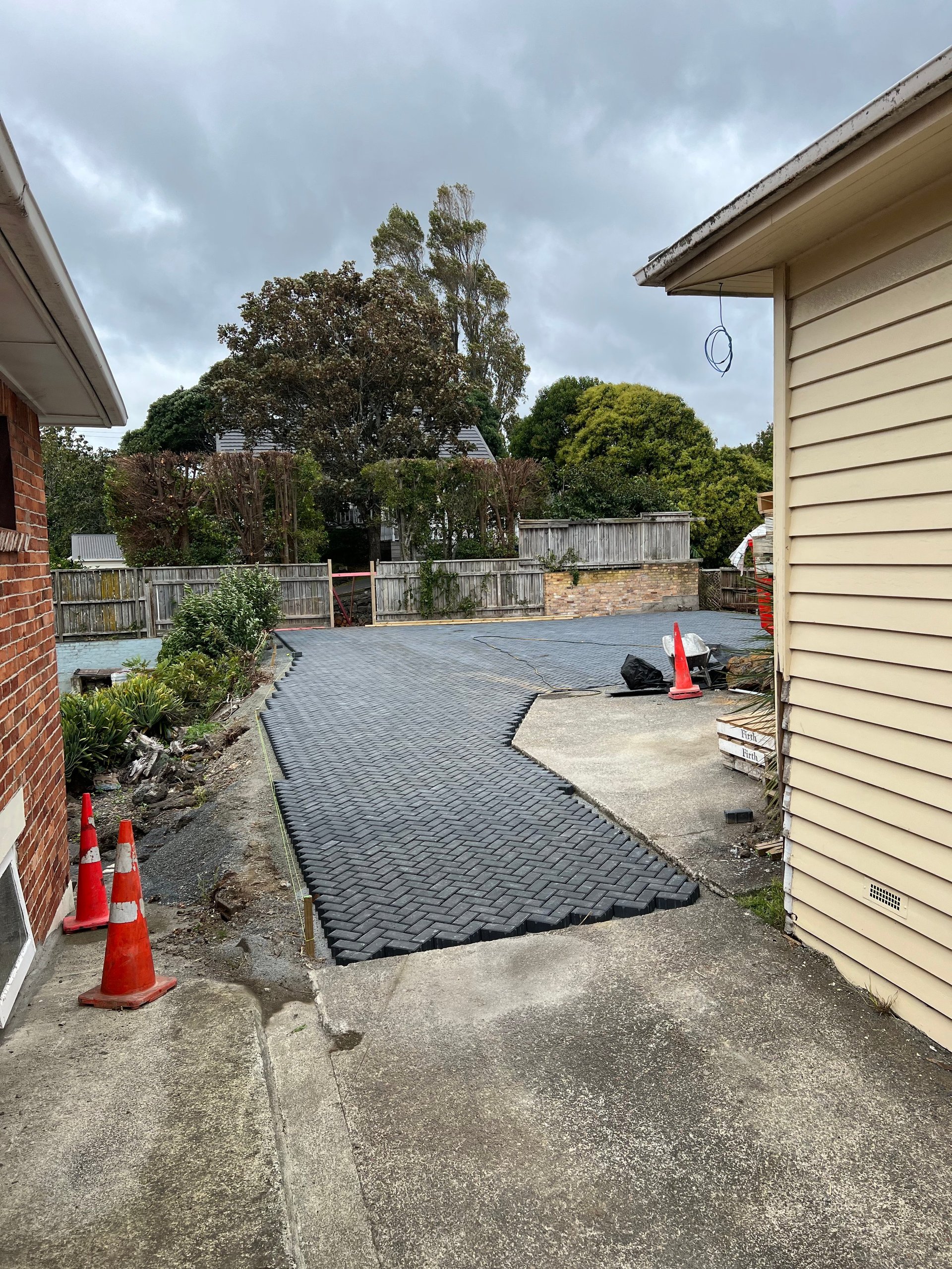 Backyard patio under construction with new paver stones, red safety cones, wooden fence, and trees visible beyond