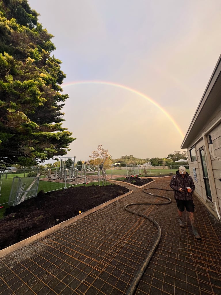 Person standing on brick patio holding a hose with a rainbow arcing across the sky above a backyard with playground equipment