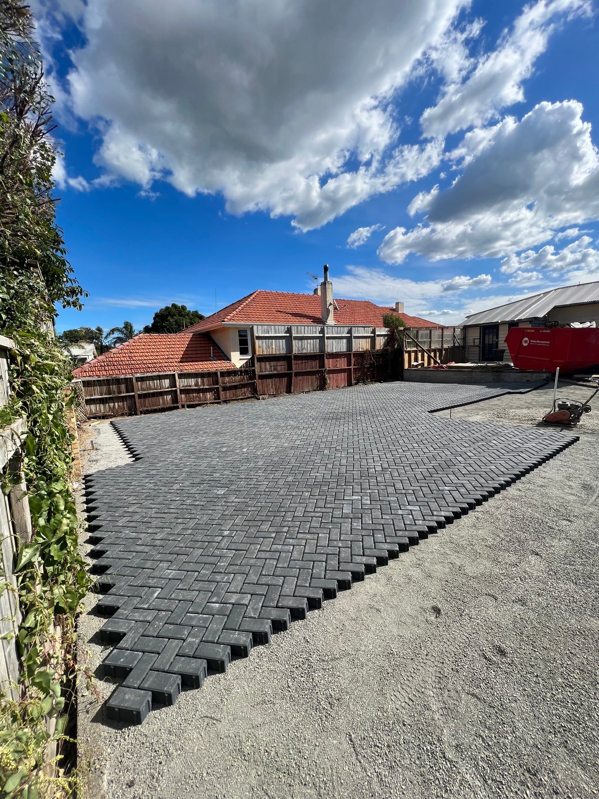 A paved driveway with a herringbone pattern leads to a rustic house with a terracotta roof and wooden gates under a blue sky with white clouds