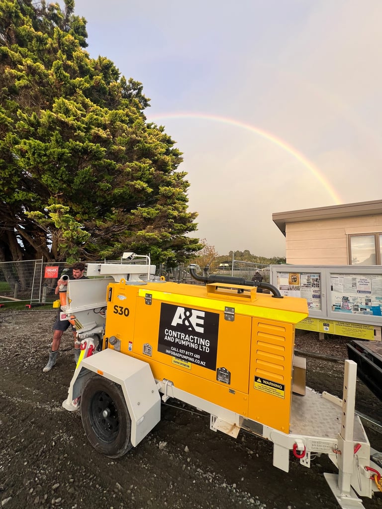 Bright yellow A&E construction equipment trailer with child standing nearby, parked near building with large tree and rainbow in sky