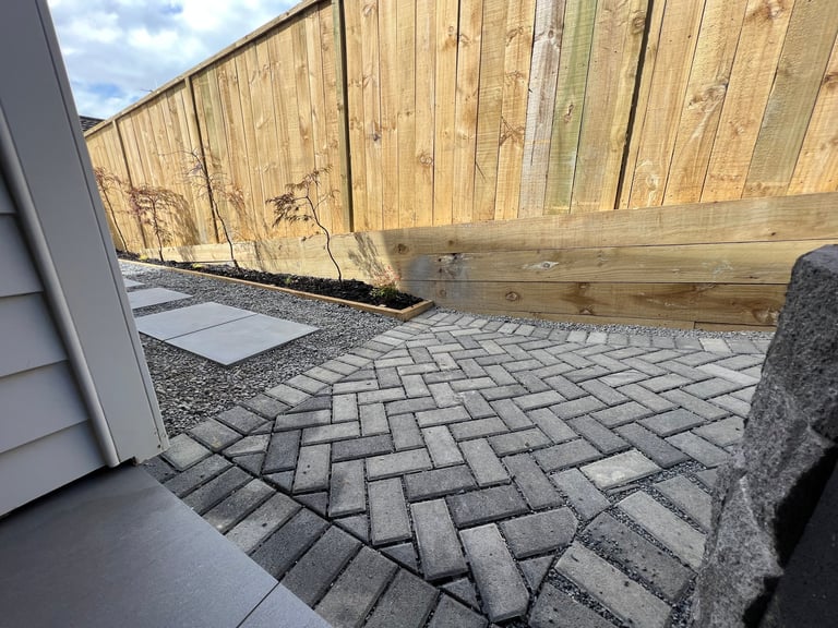 Patio with herringbone patterned pavers bordered by wooden fence and concrete foundation