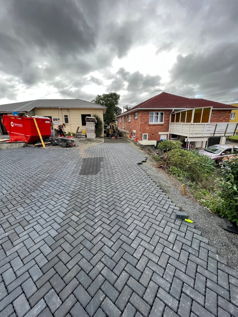 Residential driveway with paved patio, two houses with metal roofs, red equipment, and cloudy sky