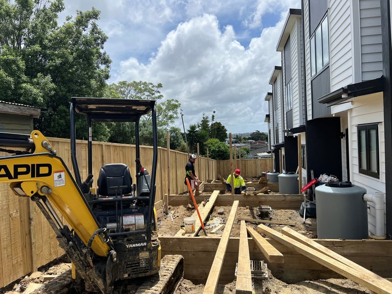 Construction site with yellow mini excavator, wooden framing materials, and workers building residential foundation next to a house