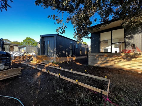 Two modern black mobile homes or tiny houses on a property with gravel ground and wooden railings, surrounded by trees and clear blue sky