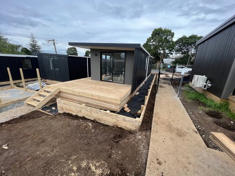 Modern black shipping container cabin with wooden deck under construction on dirt lot, surrounded by similar structures and utility poles