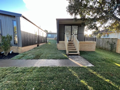 Modern prefab cabin with wooden deck and stairs surrounded by green lawn and contemporary buildings