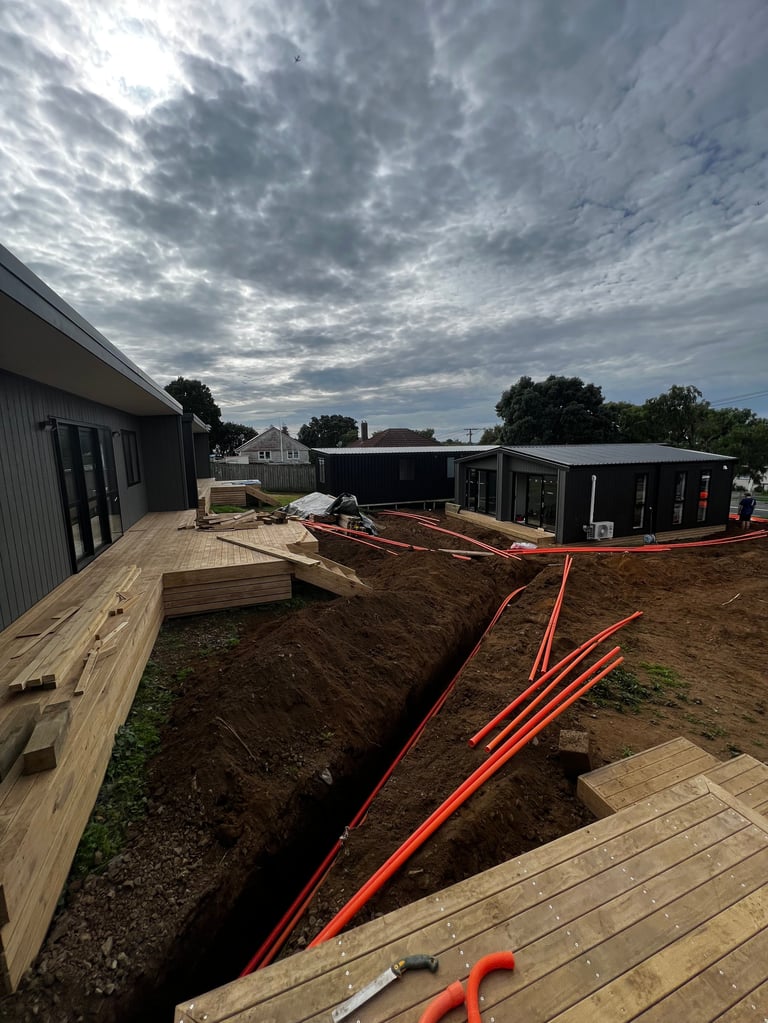 Construction site with modern buildings, red underground pipes in trenches, wooden decking, and cloudy sky
