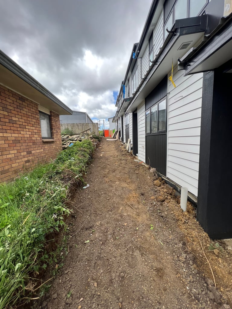 Narrow dirt pathway between brick house and white-sided building under cloudy sky