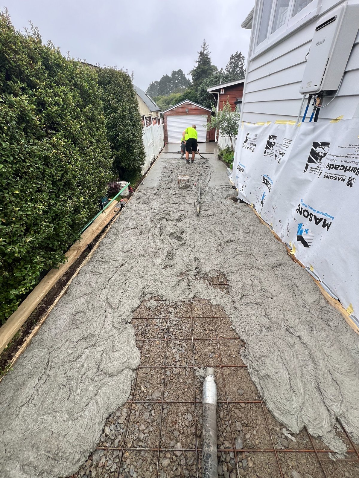 Narrow residential pathway with concrete foundation, green hedges on left, construction material on right, worker in distance